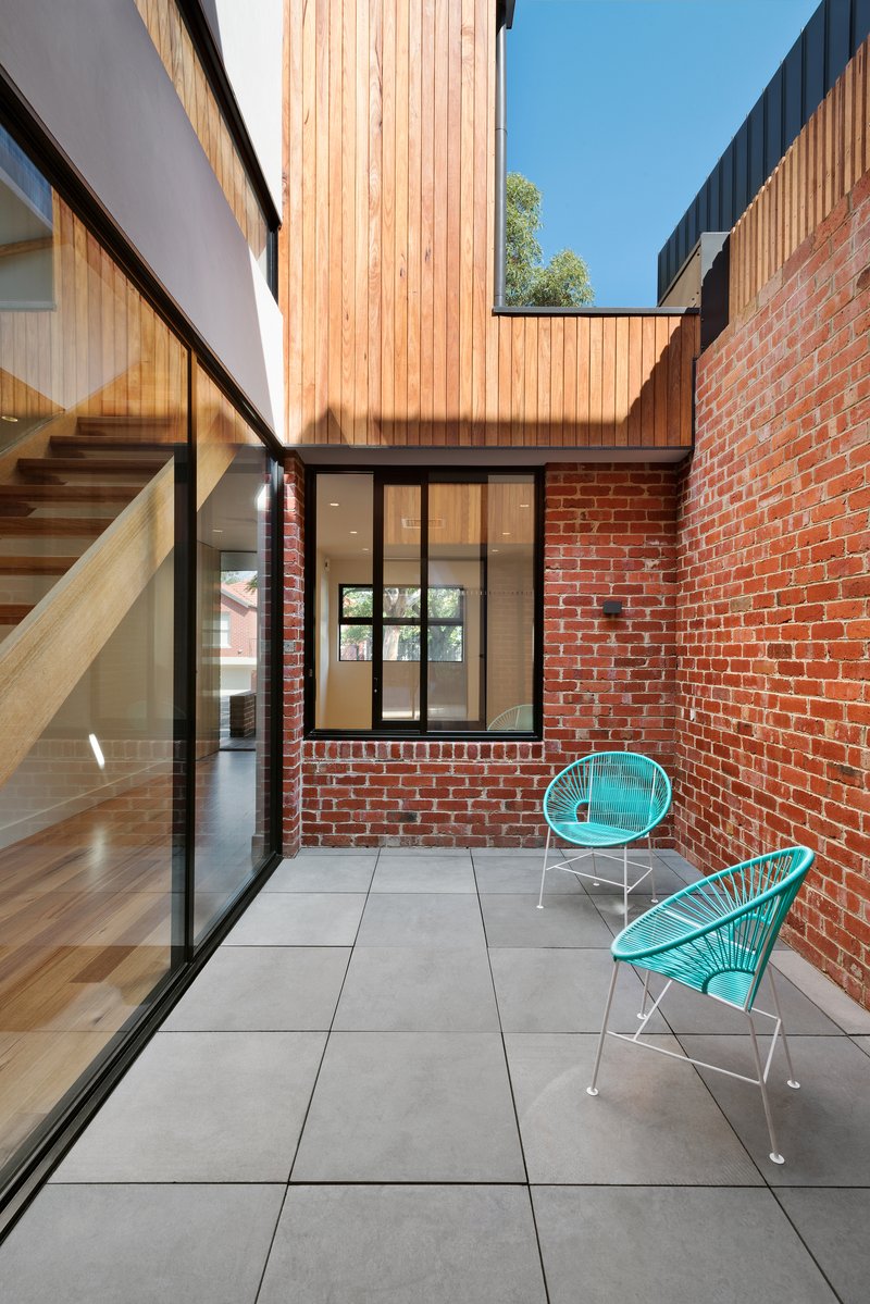 Internal courtyard with large-format grey pavers, red brick walls, and two turquoise wire chairs under blue sky
