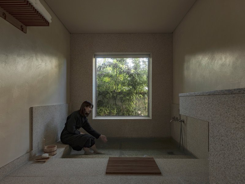 Stone-lined bathing room with a person kneeling beside the tub and window overlooking greenery