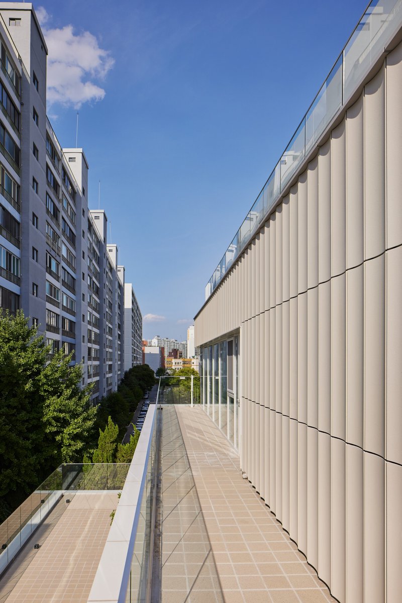 Upper terrace walkway with glass balustrade flanked by vertical metal facade and neighbouring apartment blocks