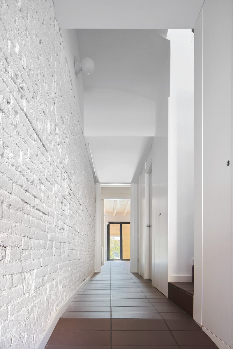Narrow corridor with painted white brick wall and terracotta tile floor leading to a distant window