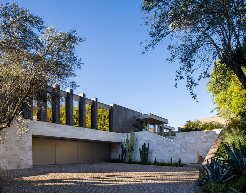 Entry facade with stone walls, timber colonnade, and garage doors framed by mature trees and cacti