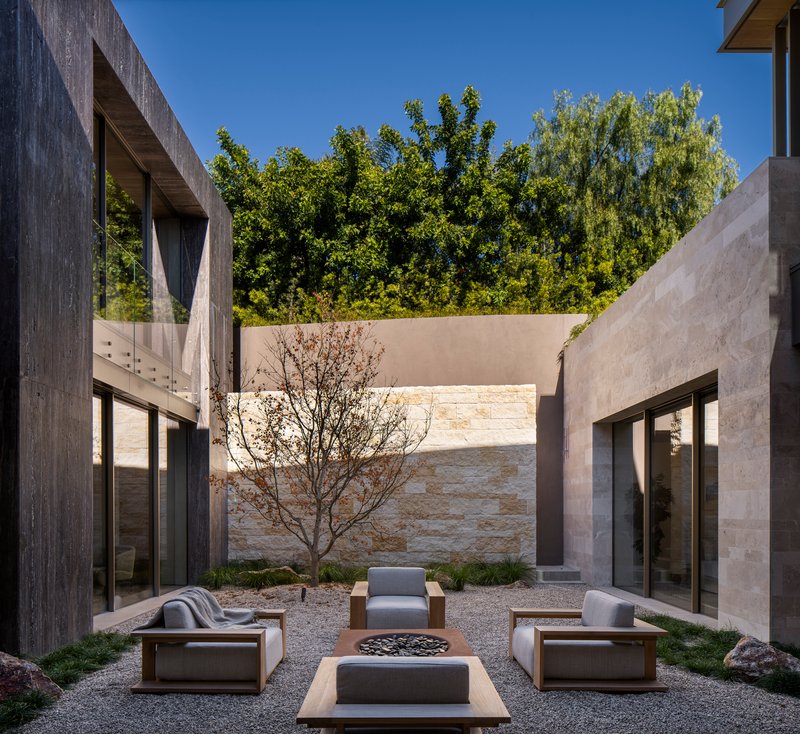 Courtyard with gravel ground plane, central fire pit, single tree, and stone walls beneath overhanging greenery