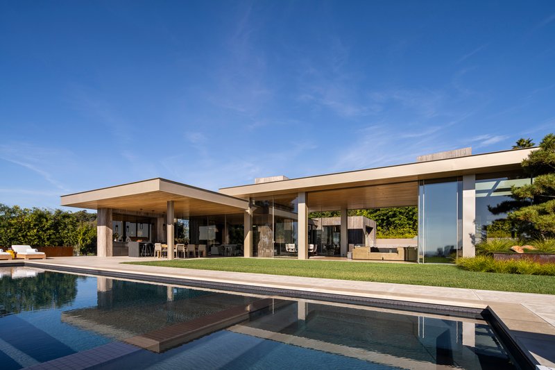 Pool terrace showing cantilevered roofs, stone columns, and reflections in still water under clear afternoon sky