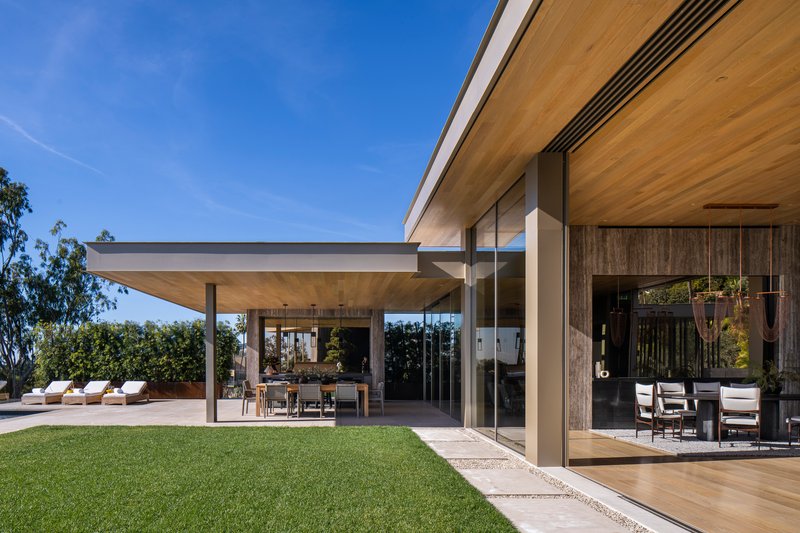 Covered terrace with timber ceiling and steel columns opening to a lawn under clear blue sky