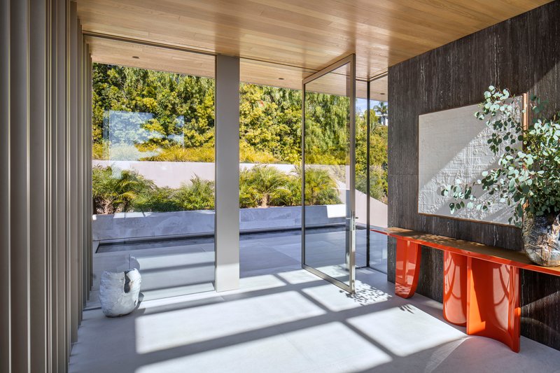 Entry foyer with glass pivot door, orange lacquered console, and view to planted terrace in sunlight