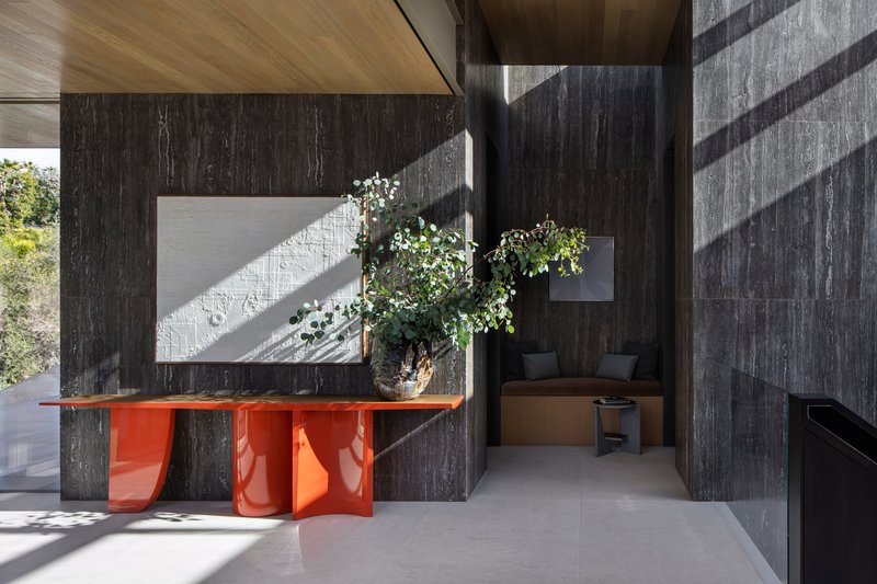 Interior view of dark vertical timber walls with an orange sculptural console table and morning sunlight streaming through openings