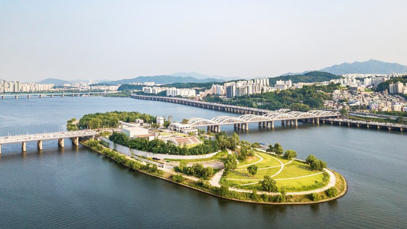 Aerial view of the landscaped island park with curved green terraces and bridges spanning the river