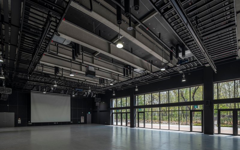 Empty multipurpose hall with exposed ceiling infrastructure and full-height glazing overlooking a wooded landscape