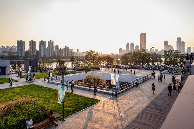 Waterfront park with lawns and sunken plaza at dusk with distant skyline reflected in the river