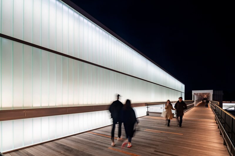 Elevated timber walkway along a backlit translucent panel wall with blurred figures at night