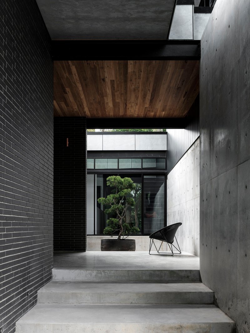 Concrete stairs ascending between black brick wall and board-formed concrete wall with timber ceiling panel overhead
