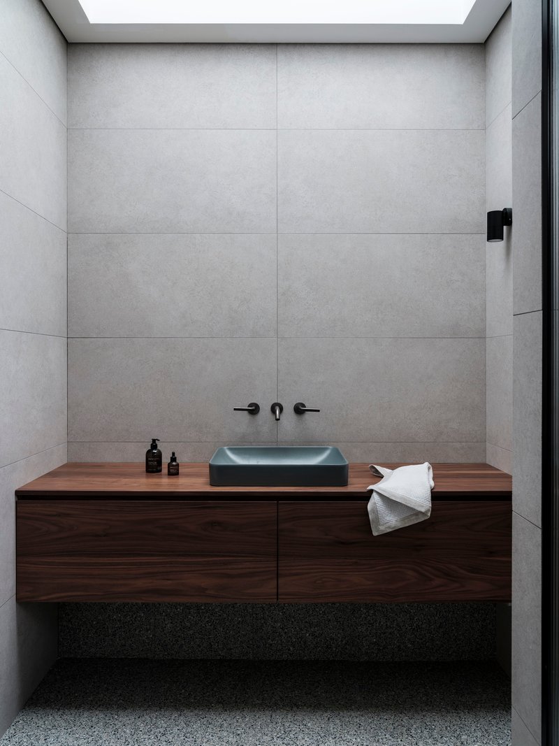 Bathroom vanity with floating walnut cabinetry and charcoal basin below large format concrete tile walls