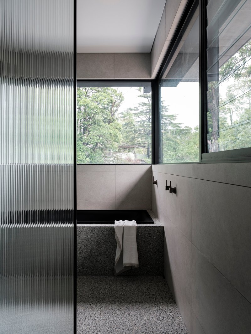 Sunken bathtub with terrazzo surround framed by fluted glass screen and windows overlooking garden