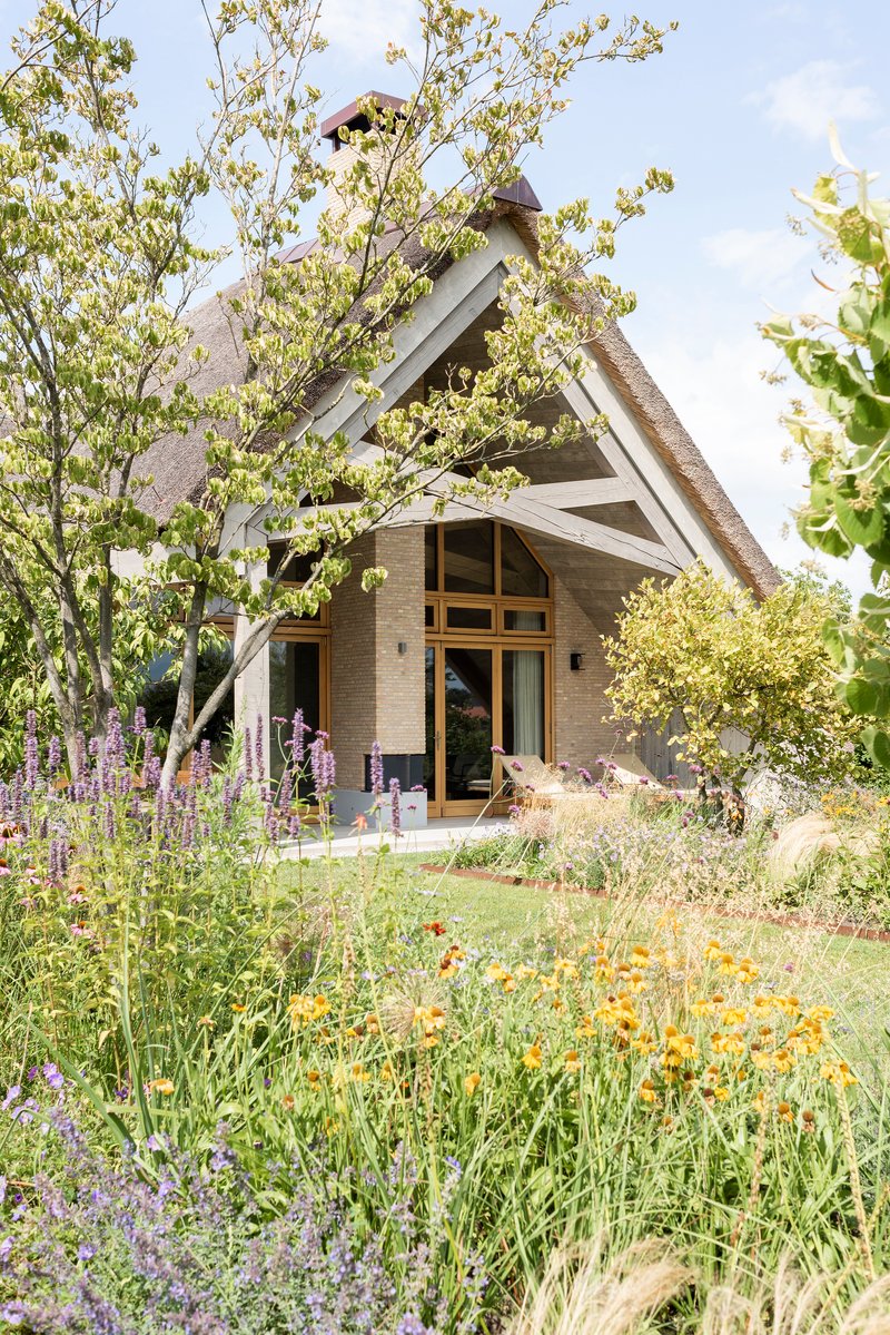Entrance facade with thatched gable roof surrounded by flowering perennials and young trees