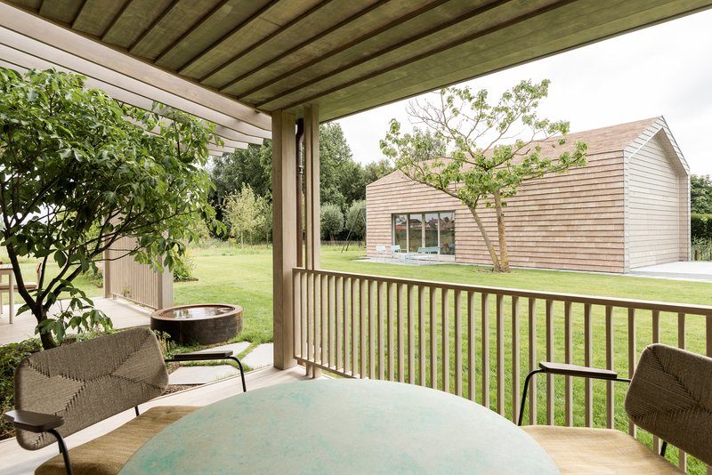 View from covered terrace through timber slat railing toward a horizontal timber-clad volume across the lawn
