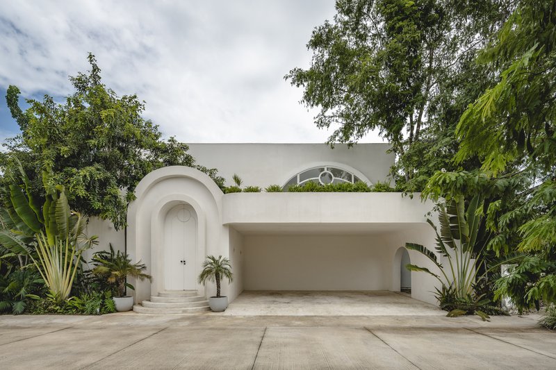 White facade with arched entrance, recessed garage, and circular window framed by banana plants and tropical trees
