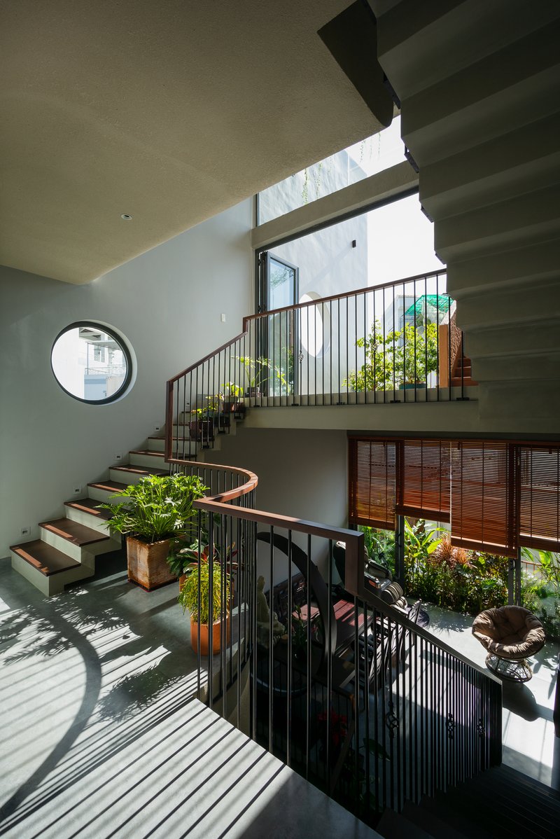 Triple-height stairwell with curved concrete steps, metal railings, and planted terraces receiving dappled sunlight from skylights