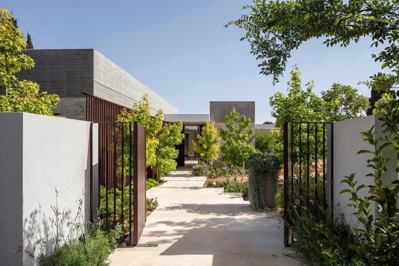 Entry pathway lined with white walls and vertical metal slats leading to concrete volumes under clear sky