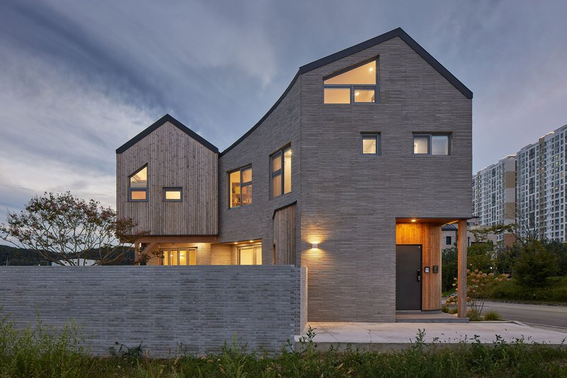 Street facade of stacked gabled volumes with dark timber cladding and illuminated windows at dusk