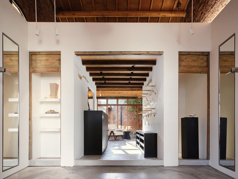 Axial view through white volumes with exposed timber beams and black display units toward a sunlit courtyard