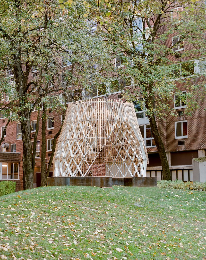 Freestanding wooden pavilion with diamond-patterned slat screen nestled among trees in a courtyard