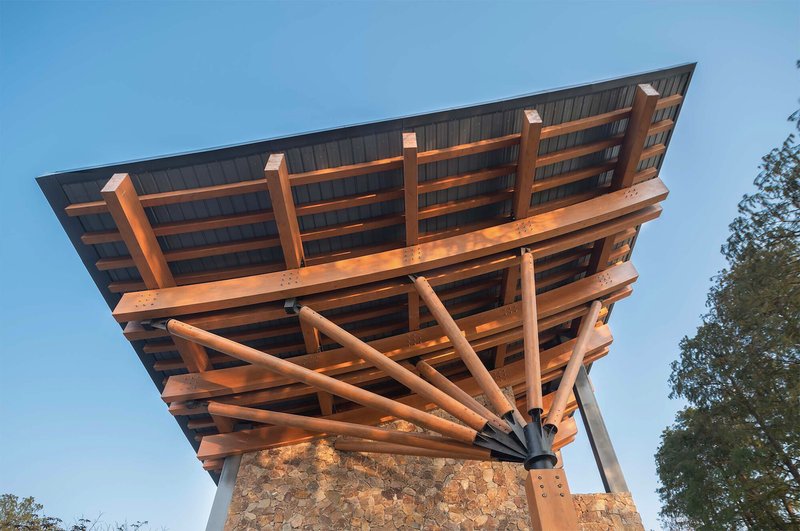 Underside view of cantilevered timber roof with radiating columns and stone base at dusk