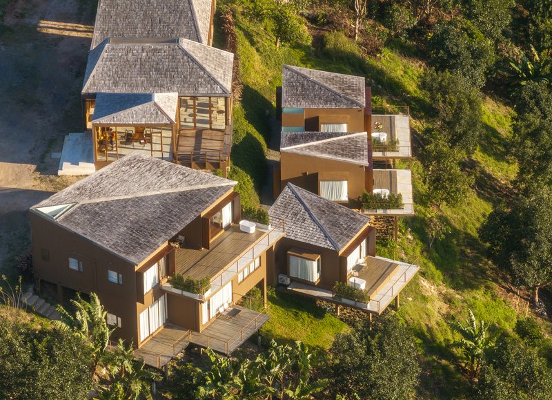 Aerial view of clustered gabled volumes with timber terraces embedded in green hillside vegetation