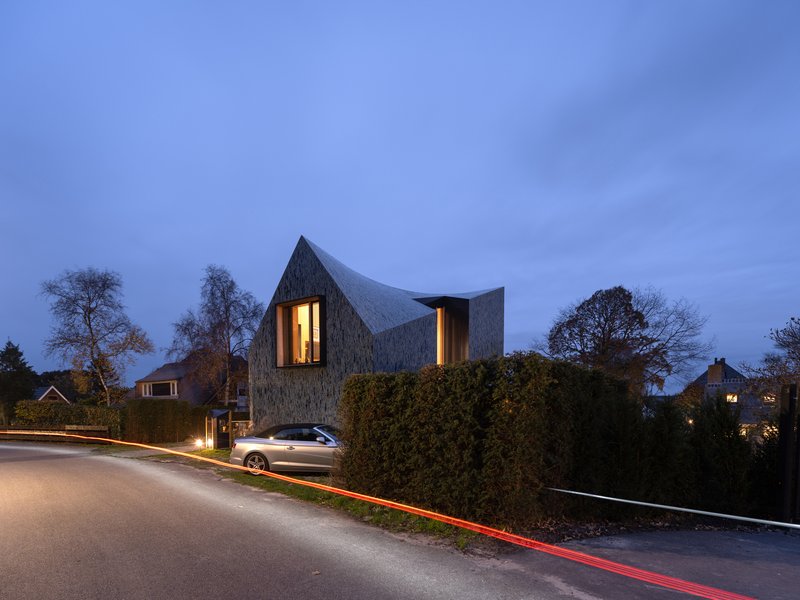 Street view of the textured facade with illuminated vertical windows at dusk behind a hedge