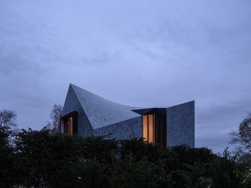 Illuminated windows glowing in the ceramic-clad volume above hedges at dusk