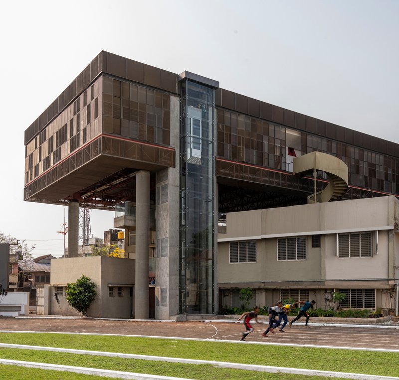 Glazed elevator shaft rising between exposed structural trusses as athletes sprint across the adjacent track