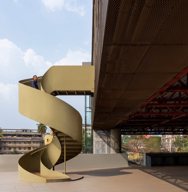 Yellow spiral staircase on a rooftop terrace with a person ascending beneath perforated metal panels