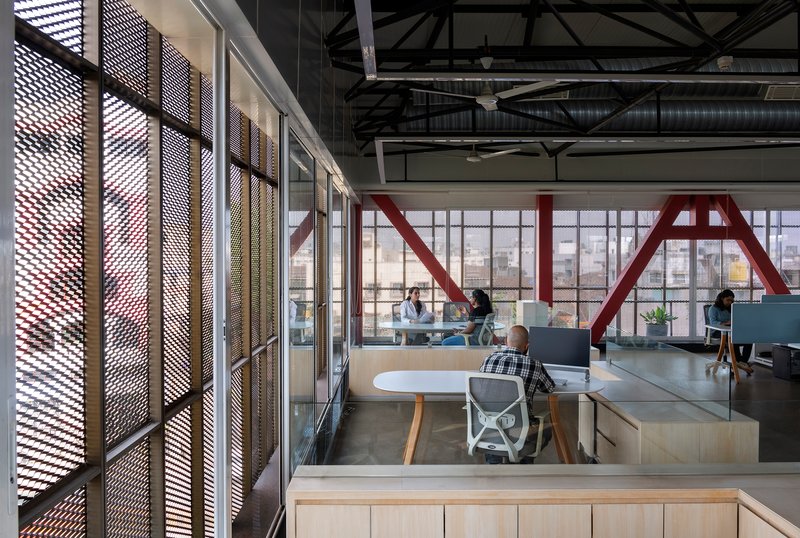 Open office space with exposed red structural trusses and perforated screen walls with workers at desks