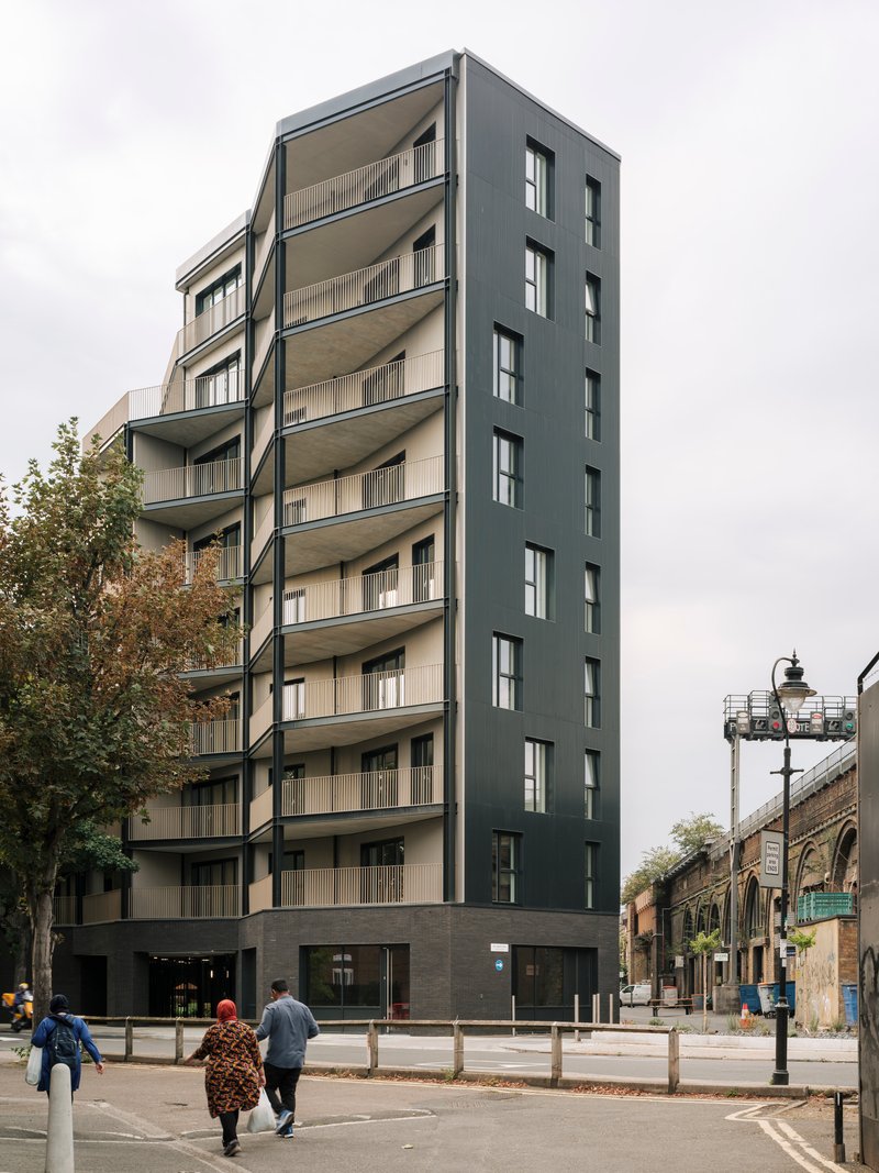 Street view of the eight-storey facade with dark cladding and recessed balconies as two pedestrians cross