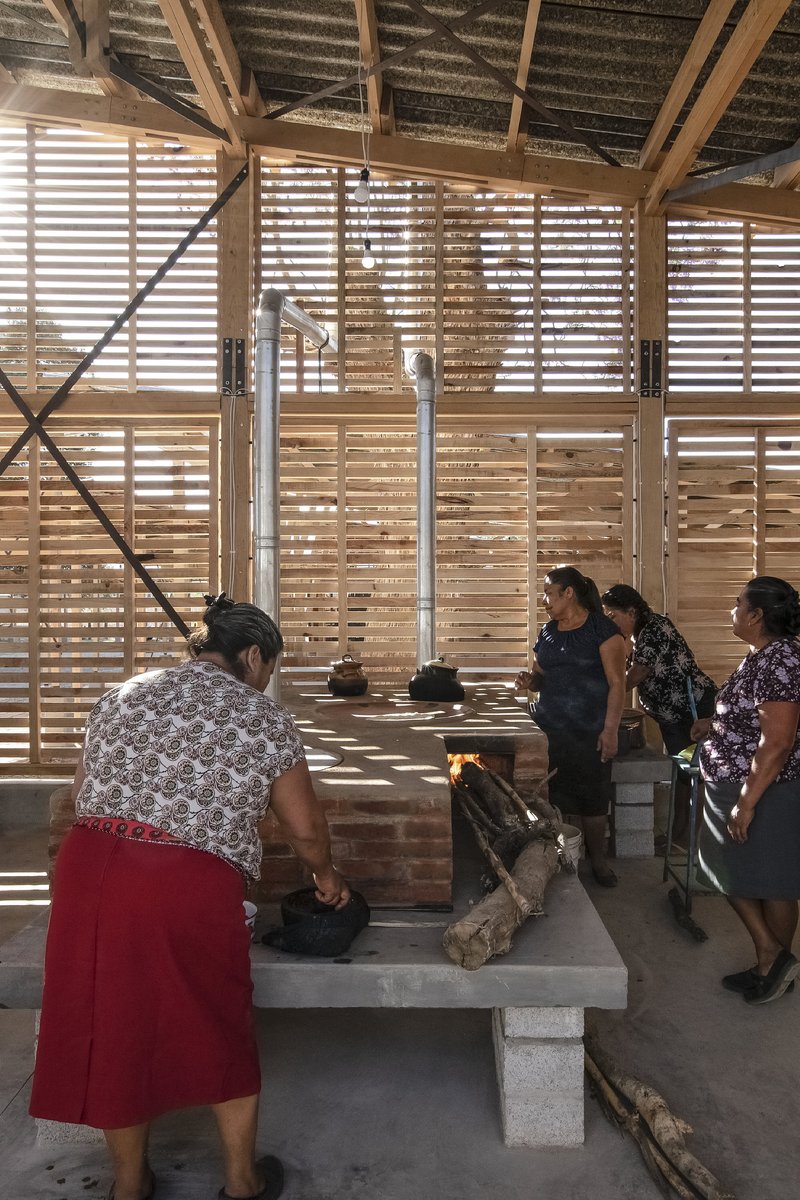 Interior view of the communal kitchen with slatted timber walls filtering dappled light onto the masonry stove