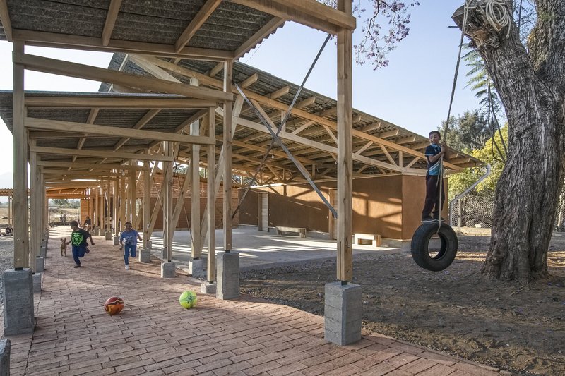 Covered outdoor space with exposed timber trusses and brick paving where children play near a tire swing