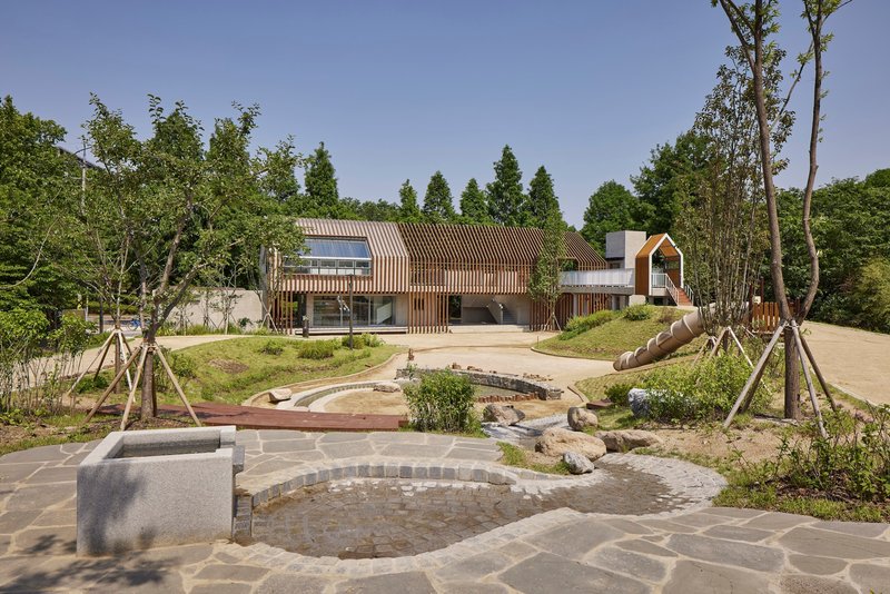 Curved stone paving and shallow water channel leading toward the timber-clad volumes under clear sky