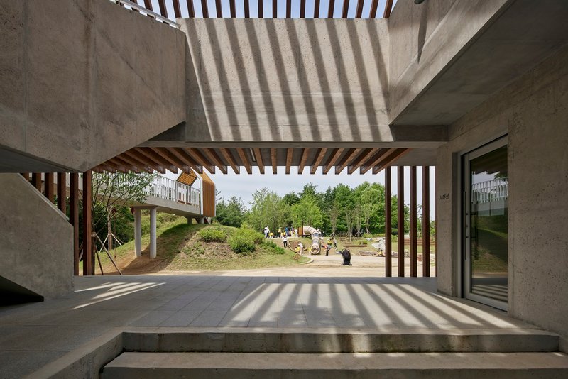 View through timber-slatted porch framing playground and wooded landscape in afternoon light