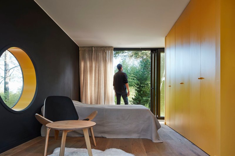 Bedroom with circular yellow-lined porthole window, yellow built-in cabinetry, and figure silhouetted in glazed doorway