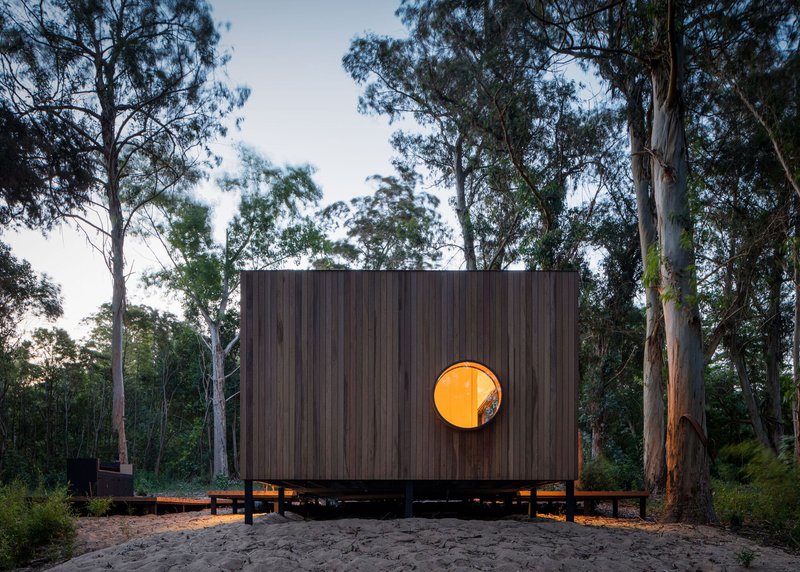 Rear facade with illuminated circular window piercing the vertical timber cladding at dusk