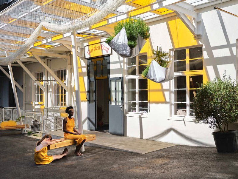 Courtyard with white tubular canopy structure, yellow-framed windows, and two figures seated on a bench in sunlight