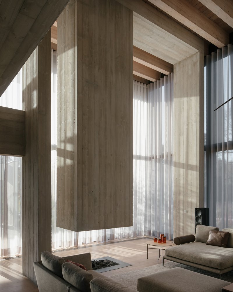 Double-height living space with floor-to-ceiling windows, timber ceiling beams and sheer curtains filtering sunlight
