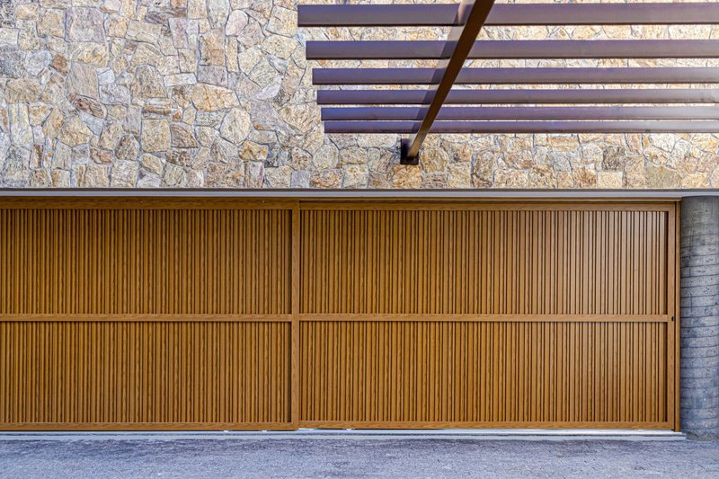 Vertical timber slat garage door beneath a slatted pergola and rough stone wall in daylight