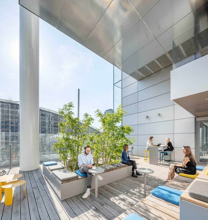 Rooftop terrace with timber decking, planted steel planter boxes and people seated under a suspended metal soffit