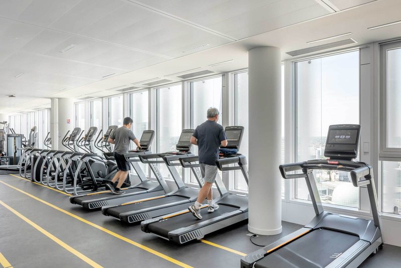 Fitness center with treadmills along the window wall and two people exercising with city views beyond