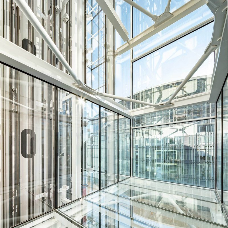 Fully glazed elevator shaft with exposed white structural bracing and glass floor panels revealing levels below