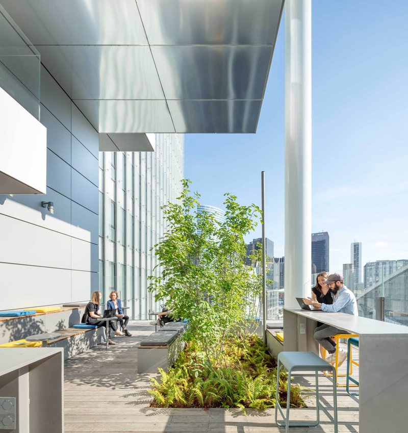Outdoor terrace with aluminum ceiling panels, planted beds, timber decking and people working at tables