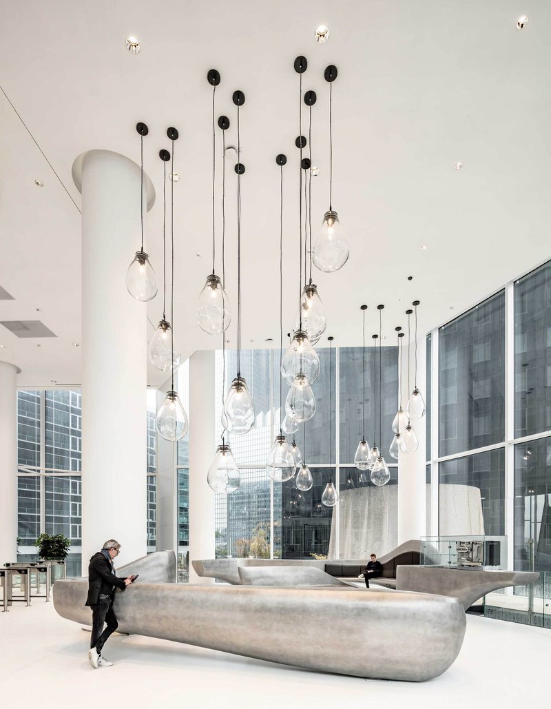 Double-height lobby with clustered pendant lights hanging above curved concrete seating and a visitor reading