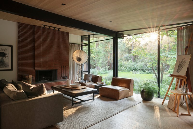 Living room with brick fireplace and corner windows framing views of trees in afternoon light