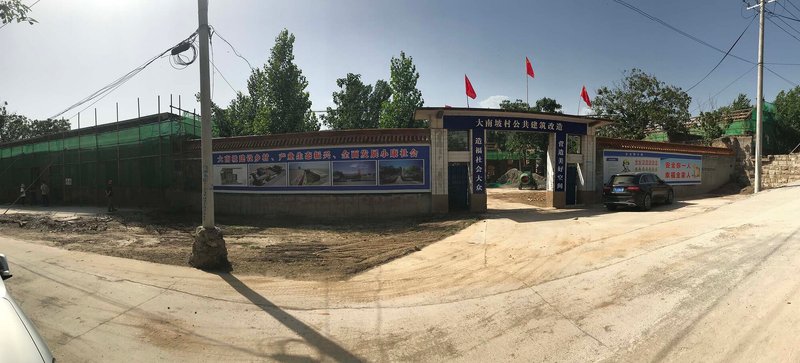 Wide-angle street view showing compound entrance with green fence and red flags under a cloudy sky