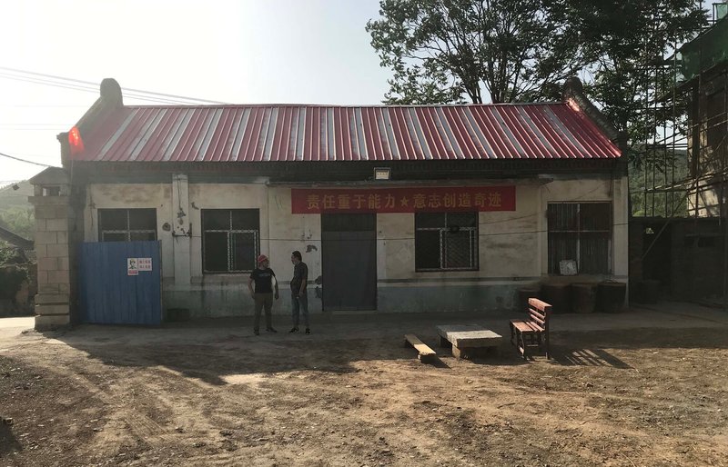 Front facade with corrugated metal roof and two figures standing in the dirt courtyard in haze